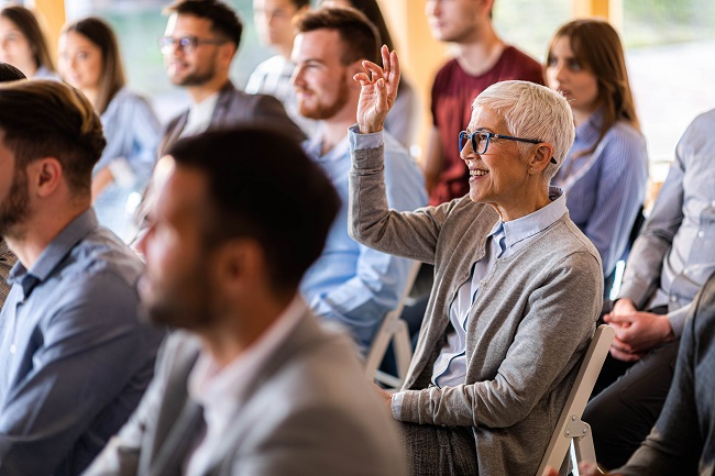 Ältere Frau, die mit Kollegen an einem Seminar teilnimmt, hebt die Hand, um eine Frage zu stellen.